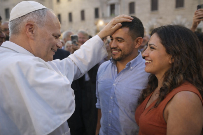 El Papa León XIV coloca su mano sobre la cabeza de un hombre latino mientras conversa con él y su pareja en una plaza, en un gesto cercano y pastoral.