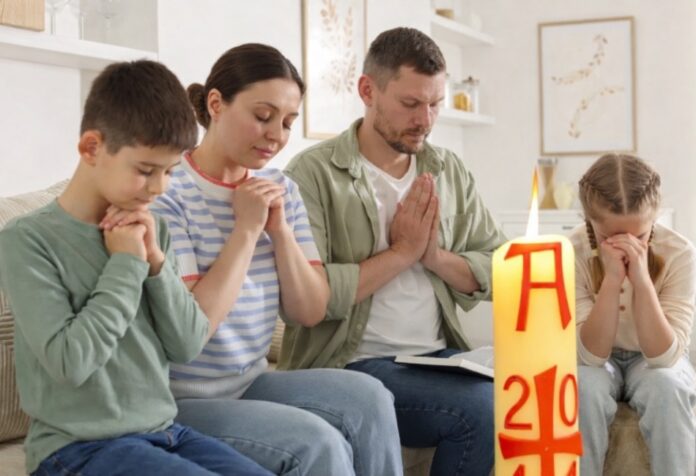Familia reunida en casa en actitud de oración, con un cirio pascual encendido en primer plano, símbolo de la Resurrección de Cristo.