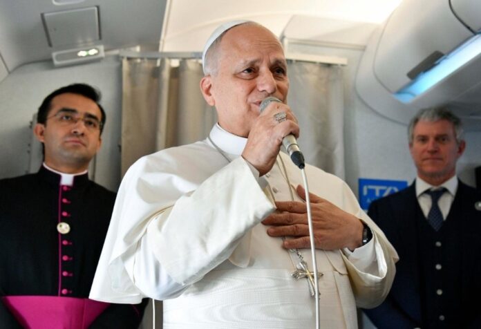 El Papa León XIV hablando con un micrófono dentro de un avión, acompañado por miembros de su equipo, durante un encuentro con periodistas.