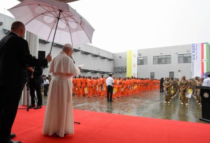 El Papa León XIV, bajo un paraguas, habla ante un grupo de reclusos formados en el patio de una prisión durante una jornada lluviosa.