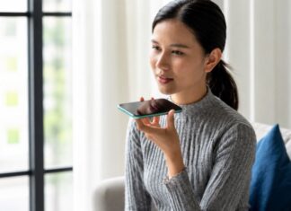 Inteligencia Artificial y pastoral juvenil: custodiar la humanidad en la era digital Joven sentada en una sala sosteniendo un teléfono móvil cerca de su rostro mientras utiliza una aplicación de inteligencia artificial por voz.