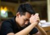 El examen de conciencia: una práctica olvidada que puede transformar nuestra vida Joven católico orando en silencio dentro de una iglesia, con las manos entrelazadas mientras realiza un examen de conciencia al finalizar el día.