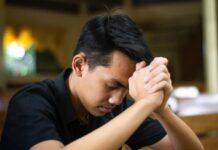 El examen de conciencia: una práctica olvidada que puede transformar nuestra vida Joven católico orando en silencio dentro de una iglesia, con las manos entrelazadas mientras realiza un examen de conciencia al finalizar el día.