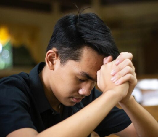 El examen de conciencia: una práctica olvidada que puede transformar nuestra vida Joven católico orando en silencio dentro de una iglesia, con las manos entrelazadas mientras realiza un examen de conciencia al finalizar el día.