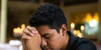¿Y si Dios sigue llamando? Joven en oración dentro de un templo católico, con las manos entrelazadas y la cabeza inclinada en actitud de discernimiento vocacional.