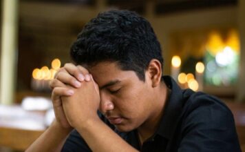¿Y si Dios sigue llamando? Joven en oración dentro de un templo católico, con las manos entrelazadas y la cabeza inclinada en actitud de discernimiento vocacional.