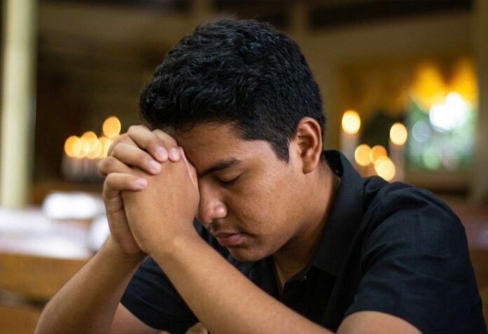 Joven en oración dentro de un templo católico, con las manos entrelazadas y la cabeza inclinada en actitud de discernimiento vocacional.