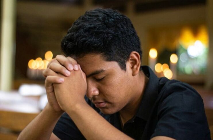 ¿Y si Dios sigue llamando? Joven en oración dentro de un templo católico, con las manos entrelazadas y la cabeza inclinada en actitud de discernimiento vocacional.